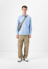 Un joven se encuentra frente a un fondo blanco, vistiendo una camisa a rayas azul y blanca, pantalones cargo beiges y un bolso gris de hombro.