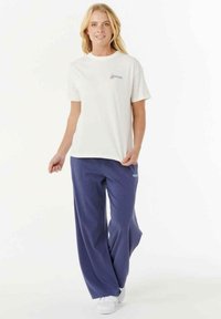 Weißes Baumwoll-T-Shirt mit Logo, kombiniert mit locker sitzenden, navyblauen Hosen aus weichem Stoff, die einen blauen Akzent am Bein haben.