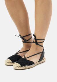 Des ballerines espadrilles noires avec un tissu tissé et une semelle en jute tressé. Liens à la cheville pour un maintien sécurisé. Bout rond avec un motif texturé.