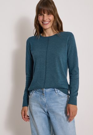 Femme souriante portant un pull turquoise à manches longues et un jean bleu clair sur un fond clair uni.