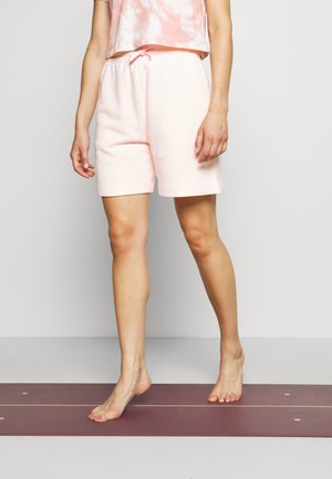 Person iført lyserøde shorts og en lyserød tie-dye top, som står barfodet på en lilla træningsmåtte mod en hvid væg.