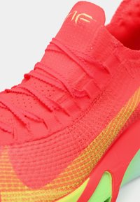 Jasnoróżowe sneakersy mają teksturowaną, oddychającą cholewkę z zielonymi akcentami. Elastyczne sznurówki i gradientowy wzór wzbogacają ich wygląd.