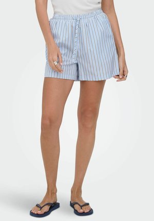 Vrouw draagt lichtblauwe en witte verticale gestreepte short met trekkoord, wit topje en donkerblauwe teenslippers, staand tegen een effen achtergrond.