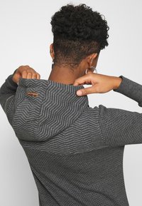 Persoon met kort krullend haar die de capuchon van een donkergrijze hoodie met kleine stippen en zigzaglijnen aanpast.