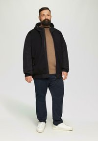 Schwarze Zip-Jacke mit Kapuze, strukturierte Einsätze; getragen über einem braunen Pullover, kombiniert mit marineblauen Hosen und weißen Turnschuhen.