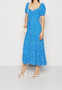 Niebieska kwiatowa sukienka maxi z bufkami, kwadratowym dekoltem i zwiewnym dołem. Połączona z białymi sandałami na koturnie i kremową torebką.