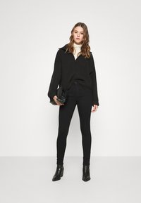 Black oversized knit sweater with a zipper, paired with black skinny jeans and black heeled ankle boots. Model holding a black clutch.
