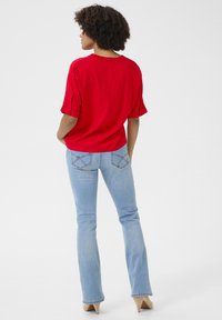 Blusa rossa a maniche corte con scollatura arricciata, abbinata a jeans a zampa d'elefante azzurri chiari con cuciture decorative sulle tasche posteriori.