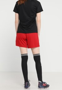 Camiseta deportiva de manga corta negra, pantalones cortos rojos, calcetas negras hasta la rodilla con rayas blancas y botas de fútbol negras con detalles.