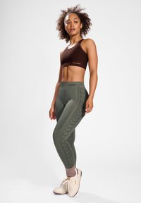Bruine cropped sporttop en olijfgroene leggings met gestructureerde patronen langs de zijkanten, gecombineerd met lichte sneakers en roze sokken.