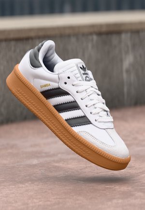Hvid Adidas Samba sneaker med sorte striber og gumisål, vist i luften over en betongoverflade og grå baggrund.