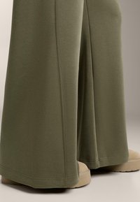 Olíva zöld anyagú, bő szabású nadrág, sima textúrával és letisztult vonalvezetéssel, világos bézs platform cipővel párosítva.