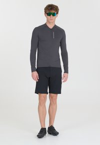 T-shirt de sport gris foncé, à manches longues avec fermeture éclair ; associé à un short noir et des chaussures de cyclisme. Équipé d'un logo réfléchissant et de lunettes de soleil vertes.