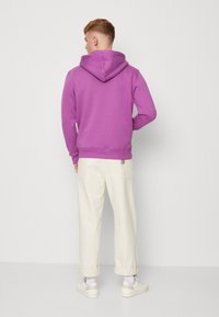 Felpa viola con cappuccio e coulisse, orli e polsini a costine, abbinata a pantaloni beige chiaro. Mostra una vestibilità rilassata e una texture morbida.