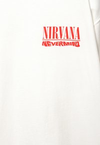 T-shirt en coton blanc avec un grand imprimé graphique rouge du texte "NIRVANA" au-dessus de "NEVERMIND" dans diverses polices stylisées.