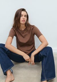 Brun bomullst-shirt med korta ärmar, rund halsringning och avslappnad passform, kombinerad med vida mörka jeans i denim med synliga sömmar.