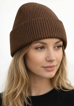 Junge Frau mit langen blonden Haaren, die eine braune gerippte Strickmütze und einen schwarzen Pullover trägt, schaut leicht nach rechts mit neutralem Gesichtsausdruck.