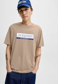 Beige bomulds T-shirt med et stort blåt og hvidt "JACK & JONES" logo på fronten, parret med mørk denim jeans.