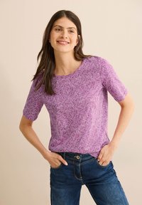 Top corto viola a maniche corte con un motivo geometrico bianco, scollatura rotonda e una texture morbida. Abbinato a jeans blu.