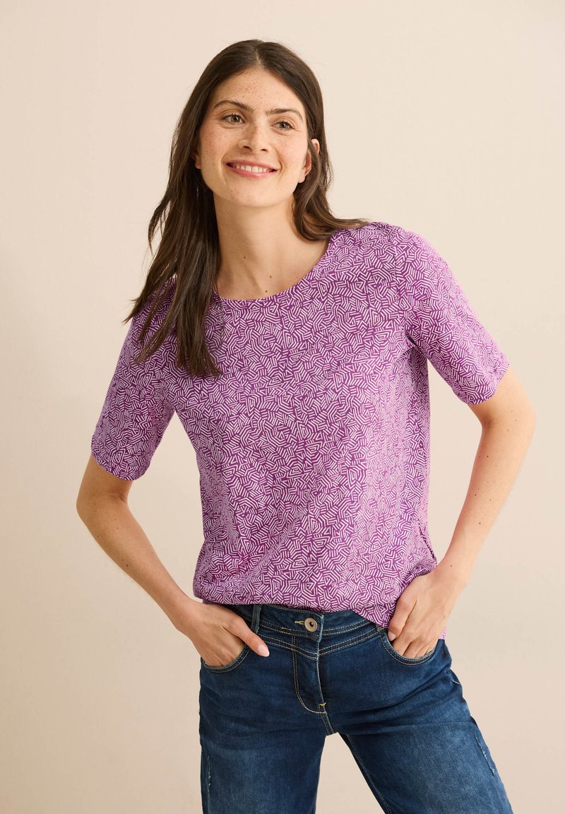 Top corto viola a maniche corte con un motivo geometrico bianco, scollatura rotonda e una texture morbida. Abbinato a jeans blu.