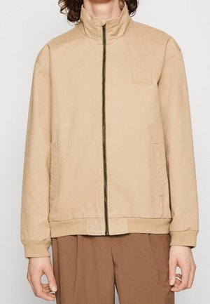 Personne portant une veste zippée beige avec poignets et ourlet côtelés, deux poches latérales et un petit logo embossé sur la poitrine.
