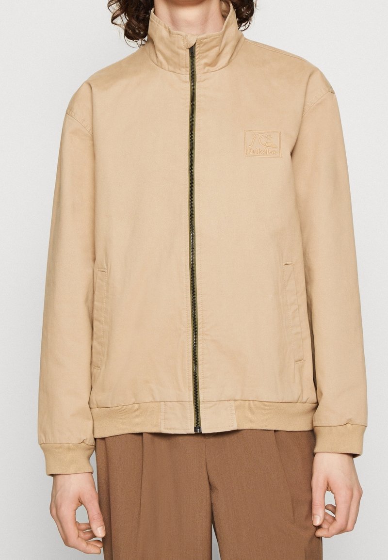 Personne portant une veste zippée beige avec poignets et ourlet côtelés, deux poches latérales et un petit logo embossé sur la poitrine.