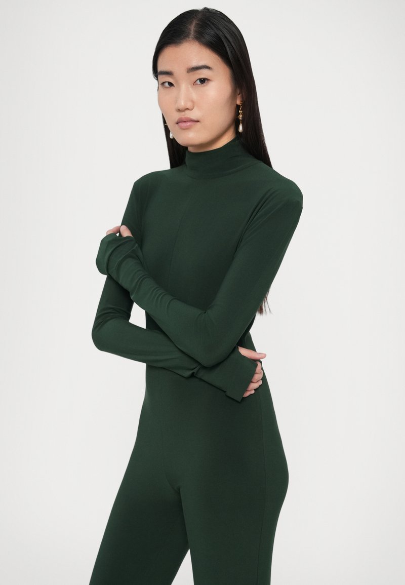 Body verde a manica lunga con collo alto, texture morbida e design aderente. Le maniche si estendono oltre i polsi per una copertura aggiuntiva.