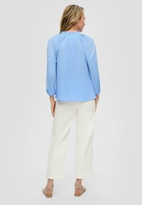 Lichtblauwe geplooide blouse met lange mouwen en gerimpelde hals, gecombineerd met crèmekleurige wijde broeken en eenvoudige sandaaltjes.