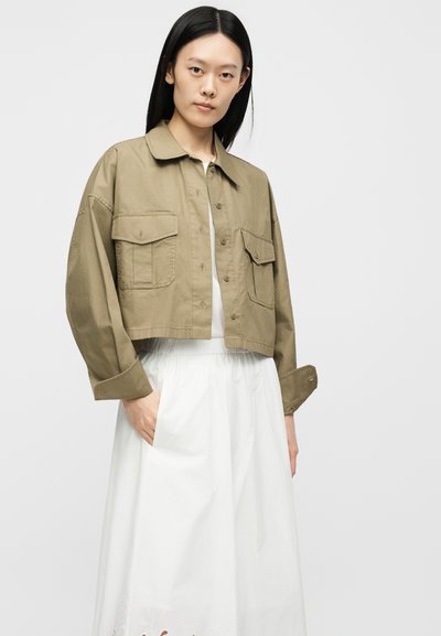 Γυναίκα με μακριά μαύρα μαλλιά που φοράει ένα κοντό μπουφάν σε ελαιόχρωμο χρώμα με κουμπιά πάνω από ένα λευκό φόρεμα, με το χέρι της στην τσέπη.