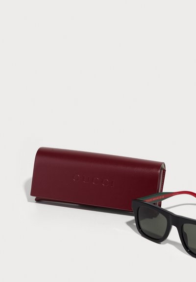 Óculos de sol pretos com lentes escuras, detalhes em vermelho e verde nas hastes. Inclui um estojo de pele bordeaux com a inscrição "GUCCI." Textura suave.