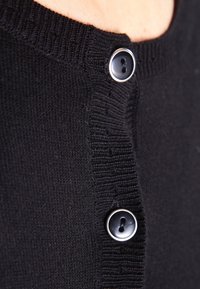 Svart stickad kofta med rund halsringning, ribbad kant och två cirkulära svarta knappar med metalliska detaljer vid öppningen.