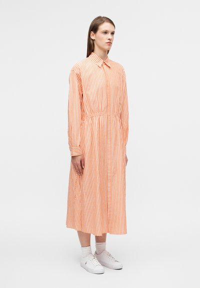 Φόρεμα με μακριά μανίκια και κουμπιά, σε πορτοκαλί και λευκές κάθετες ρίγες· διαθέτει ελαστική μέση και φτάνει μέχρι τη μέση της γάμπας. Φορεμένο με λευκά αθλητικά παπούτσια.