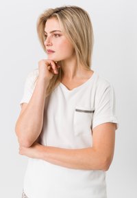 Weiße kurzärmelige Bluse mit V-Ausschnitt, die eine grau umrandete Tasche hat. Glatte Stoffqualität mit hochgerollten Ärmeln und taillenlangem Design.