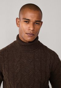 Jersey de cuello alto de punto trenzado marrón con cuello acanalado, que presenta patrones texturizados y un diseño ajustado. Material suave y grueso.