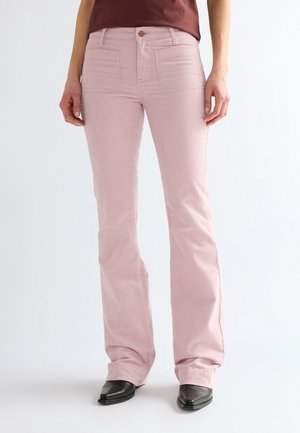 Personne portant un pantalon en velours côtelé rose clair et des chaussures noires pointues, debout sur un fond blanc.