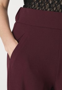 Bordowe spodnie z wysokim stanem o gładkiej fakturze, z bocznymi kieszeniami i płaskim pasem, prezentujące minimalizm w detalu.