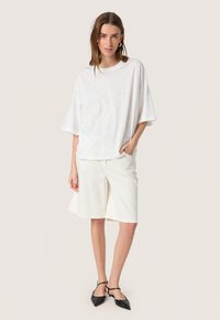 Hvid oversized t-shirt med spredte udsmykninger, parret med cremefarvede shorts og sorte flade sandaler med stropper. Ensfarvet baggrund.