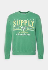 Zelená mikina z bavlny s kulatým výstřihem, na které je žlutý nápis "VINTAGE SUPPLY CENTRAL DIVISION" a "SUPER BOWL Champions."