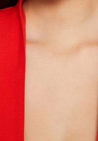 Czerwony materiał o gładkiej fakturze, pokazujący część dekoltu na tle jasnej karnacji. Brak widocznych wzorów lub akcentów.