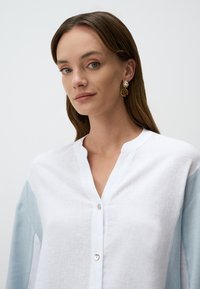 Camicia button-up bianca e azzurro chiaro con scollo a V, maniche lunghe e tessuto testurizzato. Presenta una chiusura con un solo bottone al collo.