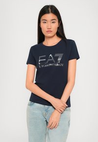 Mørkeblå kortermet t-skjorte med sølvfarget logo-design "EA7 Emporio Armani." Stoffet ser glatt og bomullsliknende ut, med rund hals.