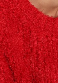 Pull en tricot rouge duveteux présentant une surface texturée et un large décolleté. Les fibres créent une apparence douce et moelleuse.