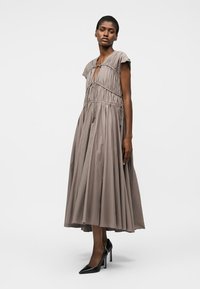 Žena oblečená v taupe plisovaných šatách midi dĺžky s krátkymi rukávmi a čiernymi vysokými podpätkami, stojaca na jednoduchom pozadí.