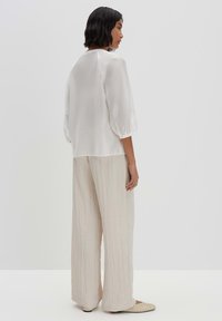 Blusa bianca leggera con maniche a tre quarti, abbinata a pantaloni beige larghi e sgualciti e ballerine beige, vista da dietro.