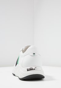 Białe sneakersy z czarnymi akcentami, zielonymi paskami i teksturowanym tyłem. Posiadają błyszczącą srebrną płytkę z logo oraz grubą, amortyzującą podeszwę.