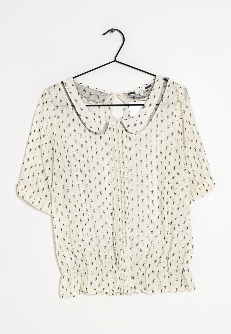 Blouse blanche avec petit motif noir, col Peter Pan, manches mi-longues, dos avec ouverture en forme de goutte, et ourlet froncé, suspendue à un cintre noir sur fond blanc.