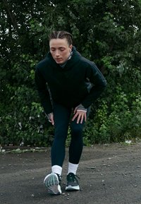 Oseba v črni jakni in legginah raztegne tele na gravelni poti s gostim zelenim rastlinjem v ozadju.