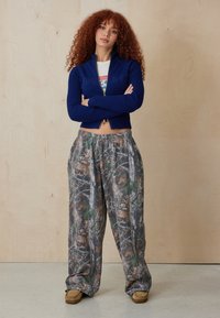 Maglione zippato a coste di colore blu navy con colletto alto, abbinato a pantaloni sportivi larghi con motivo mimetico caratterizzati da tonalità terrose e motivi di alberi. Calzature marroni.