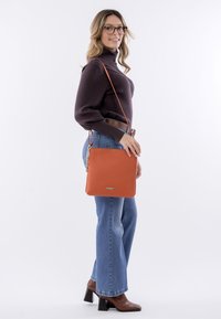 Orange leather handbag with a smooth texture, square shape, and discreet logo. Features a long strap for crossbody wear.
