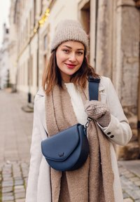 Námořnická kožená crossbody taška s kulatým klopou, spárovaná s béžovým pleteným šálou a rukavicemi, nošena přes světle svetr a kabát.